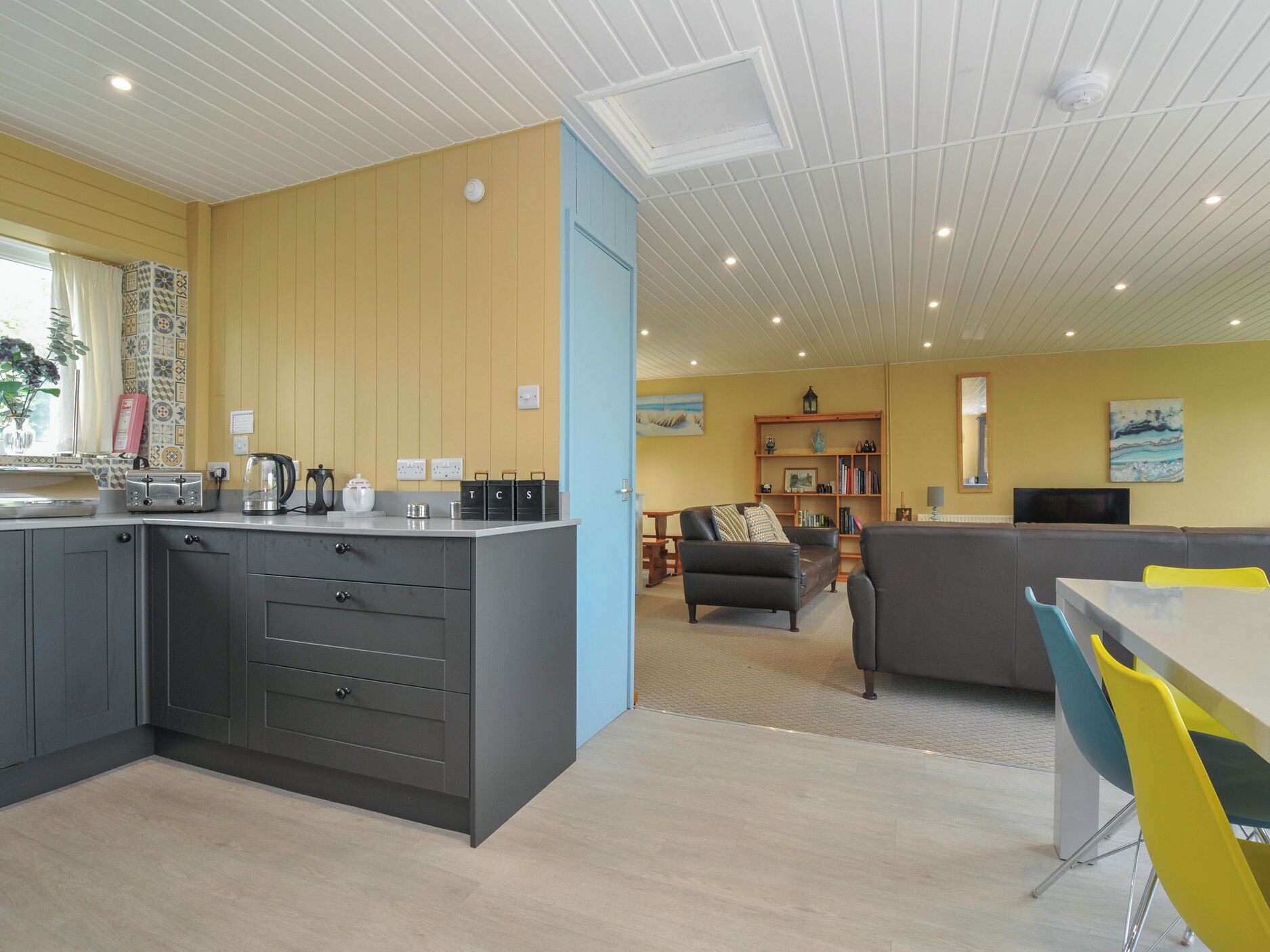 Una cocina moderna con armarios grises y una isla central, una sala de estar con un sofá y una mesa de centro, y un comedor con una mesa amarilla y sillas azules.