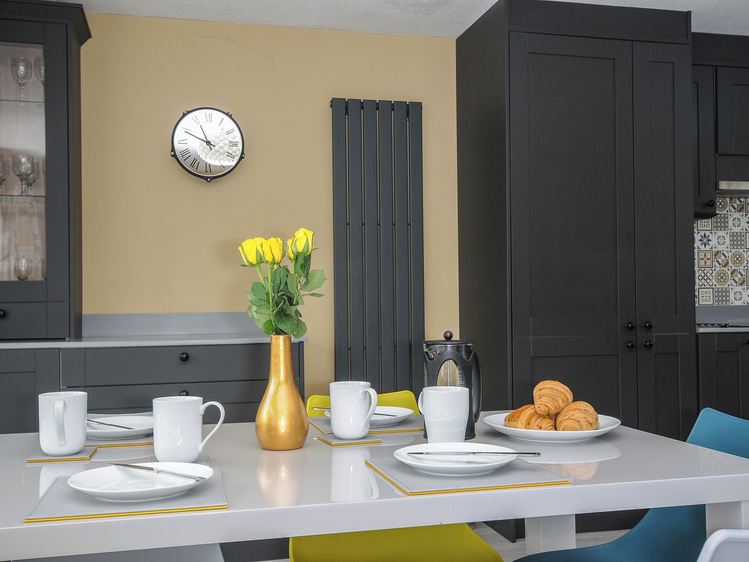 A modern kitchen with a white table set for breakfast, a clock on the wall, and a radiator.