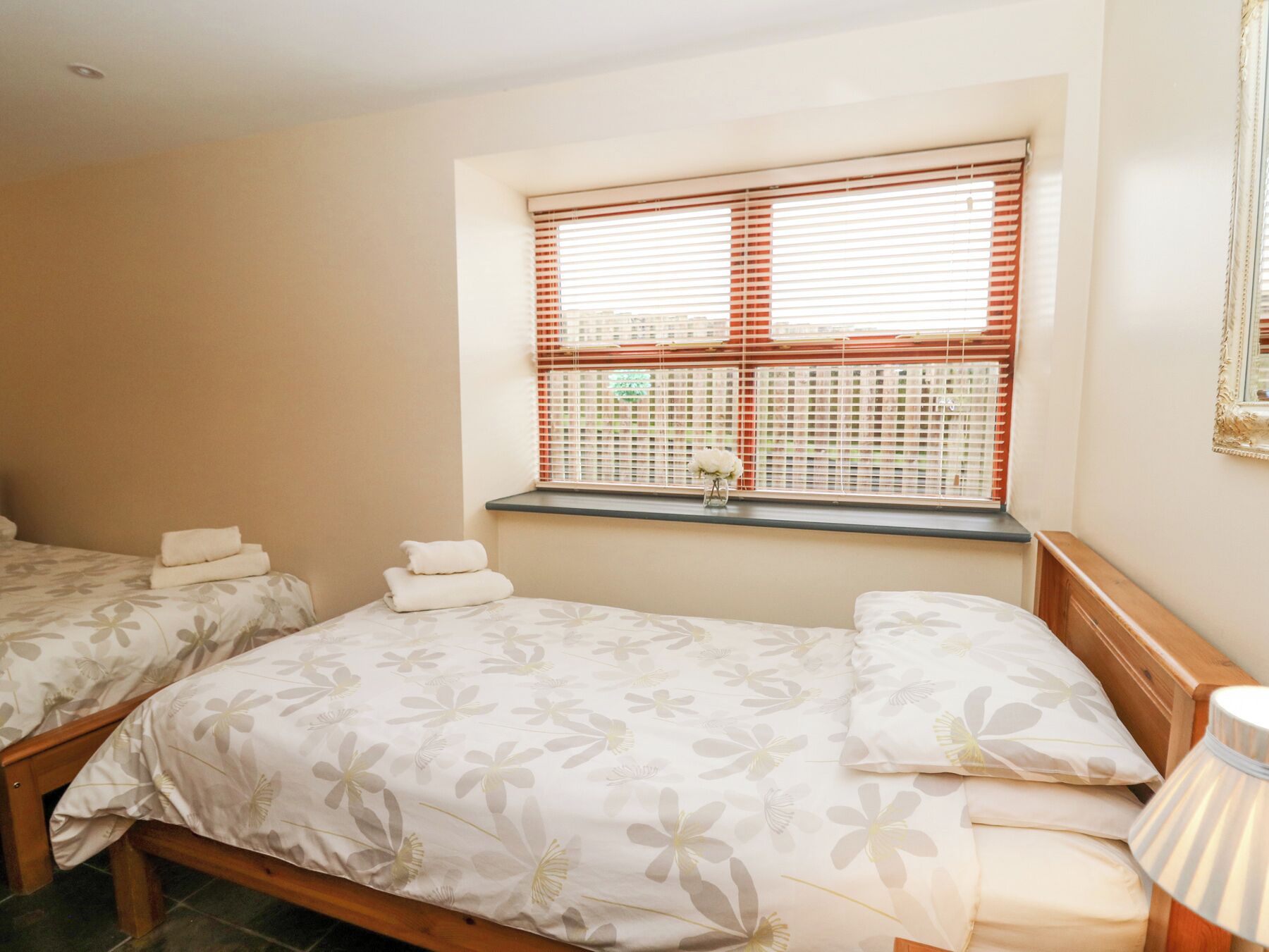 A hotel room with two beds, a window with blinds, a small table, and a lamp.
