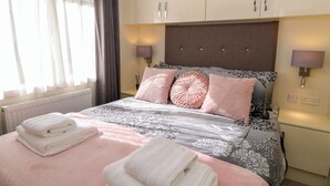 Cama bem arrumada com almofadas e toalhas, cabeceira com luminárias fixadas na parede e janela com cortinas.