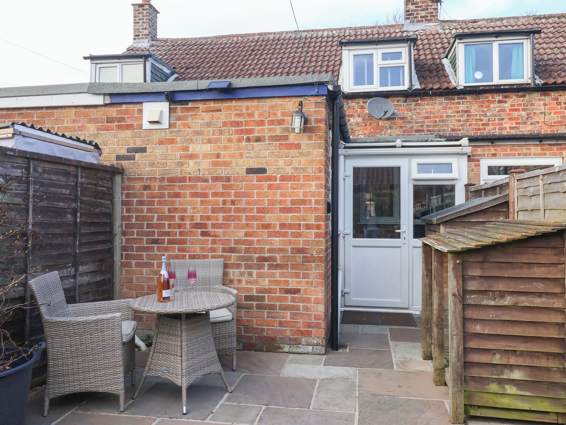 A brick house with a tiled roof, a white door, and a small patio with wicker furniture and a table.