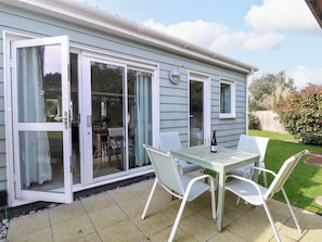 A patio with a table and chairs, a bottle of wine, and a sliding glass door leading to an indoor area with a dining table and chairs. - Plimoth N9 (Yarmouth)
