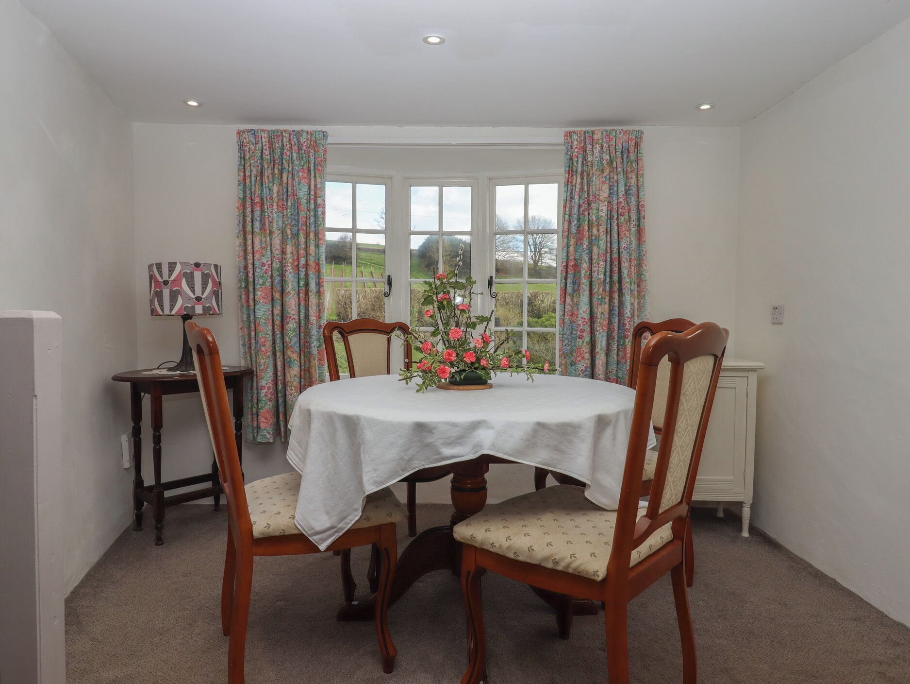 A dining area with a round table, four wooden chairs, a floral centerpiece, and a window with curtains.