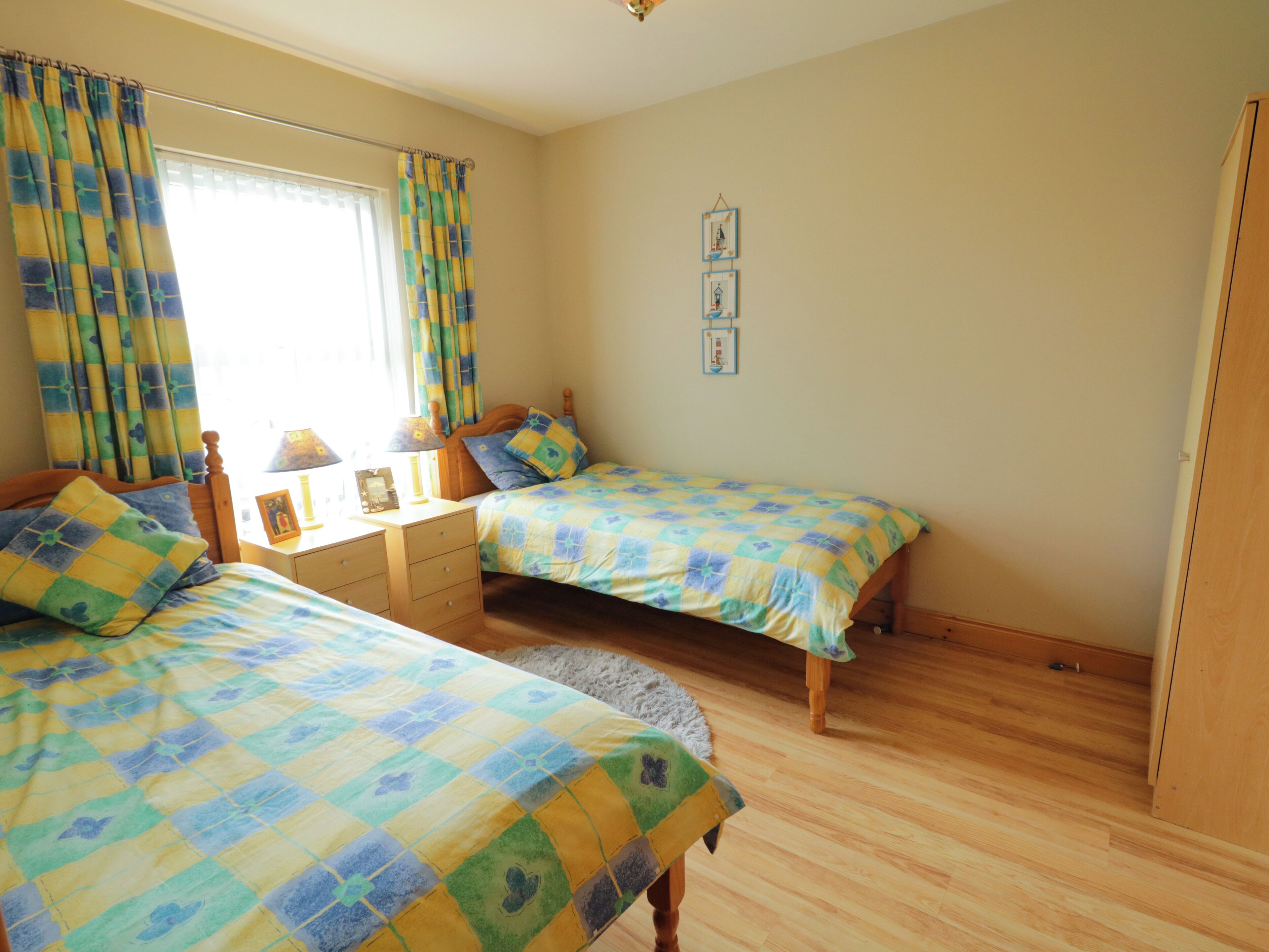 A bedroom with two single beds, a wooden dresser, a nightstand, a lamp, and a window with curtains.