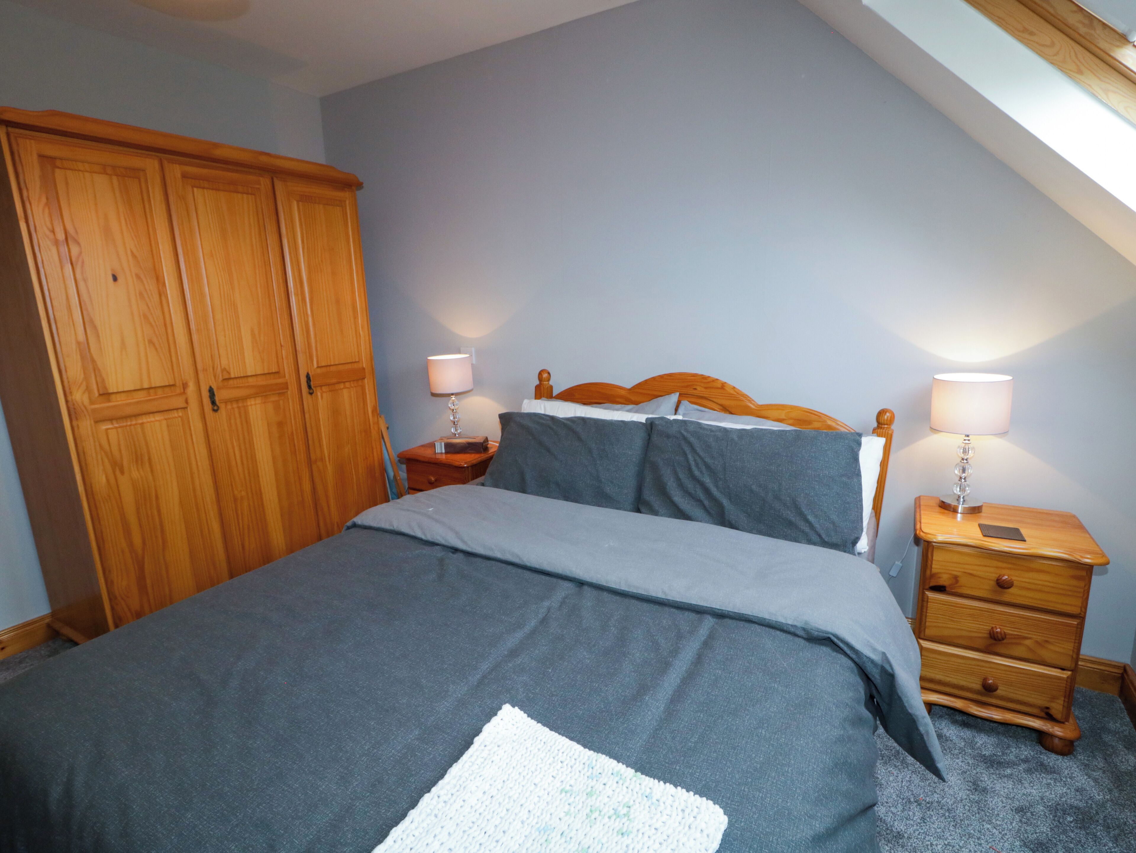 A bedroom with a bed, wooden wardrobe, two bedside tables with lamps, and a sloped ceiling.