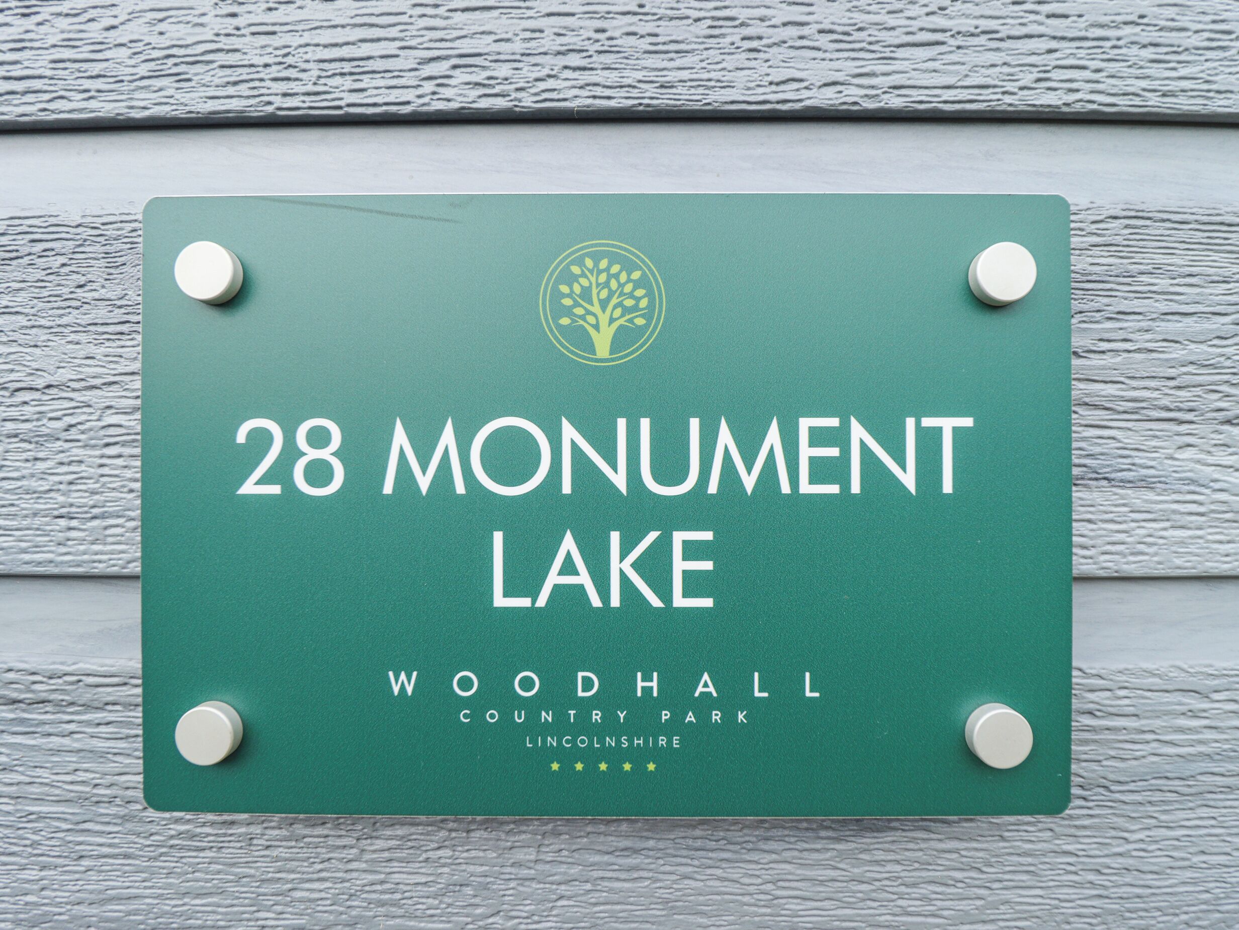 Segnaletica verde con testo bianco che indica "28 Monument Lake Woodhall Country Park Lincolnshire" e un logo raffigurante un albero.
