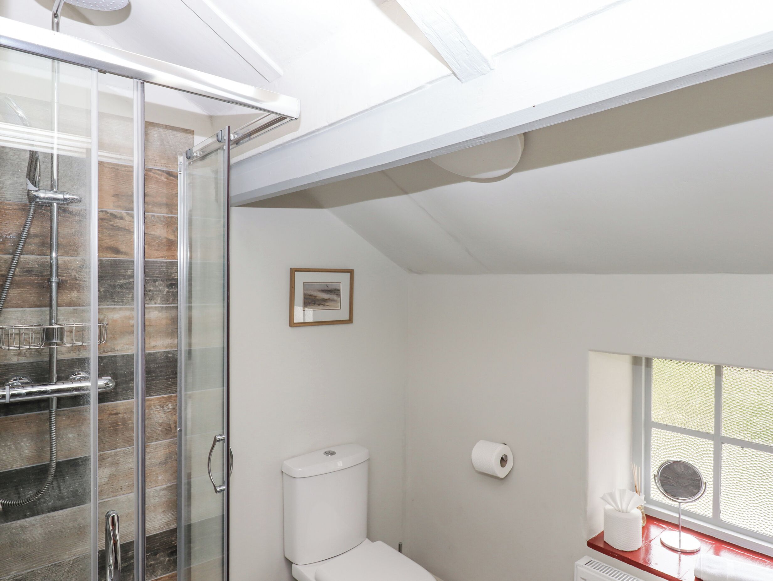A modern bathroom with a glass shower enclosure, a toilet, a framed picture on the wall, and a window with a view.