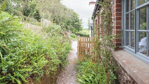 A narrow pathway flanked by dense foliage and a wooden fence, leading to a brick building with a window. - Hole Farm Stables (Newton Abbot)