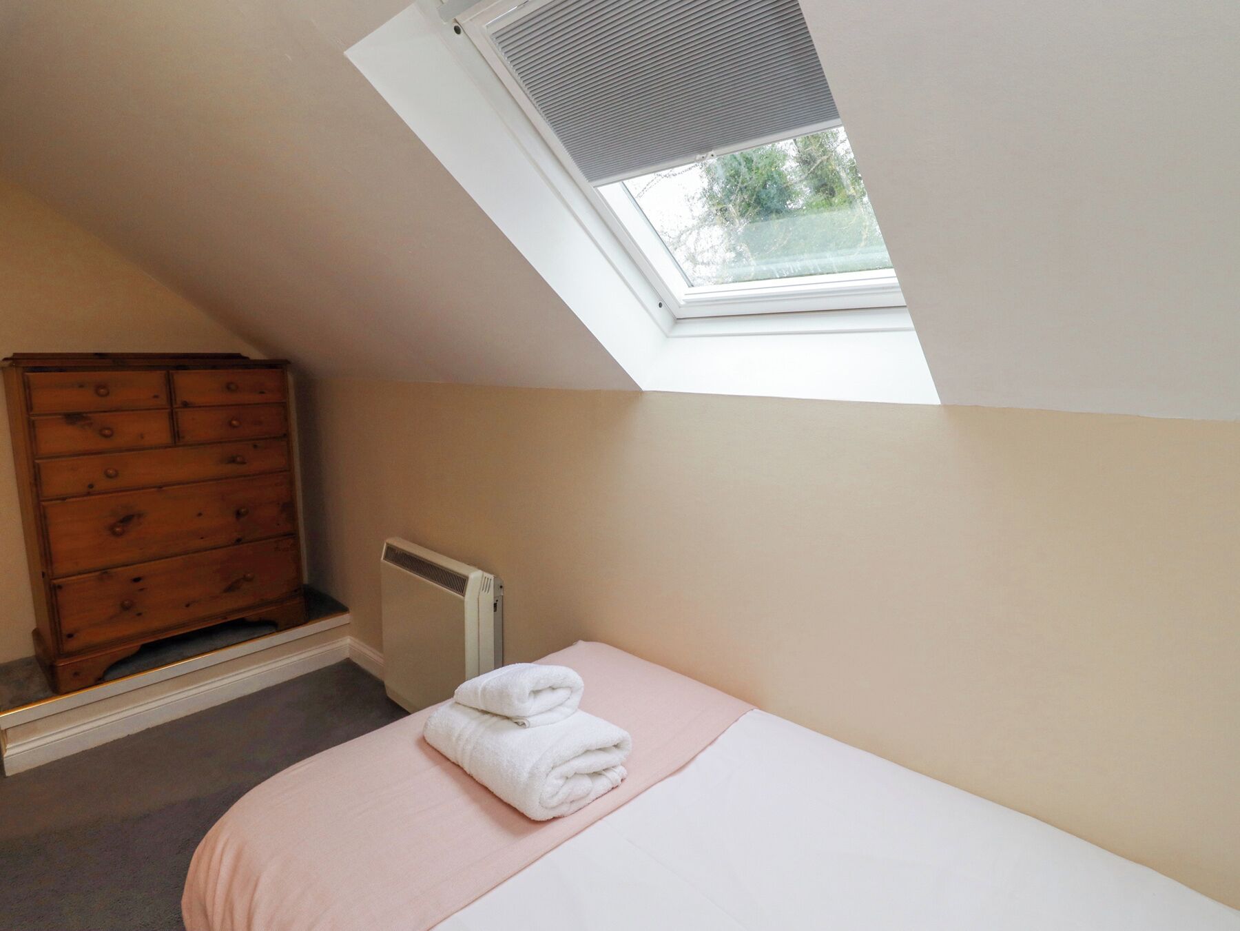 A bedroom with a sloped ceiling, a skylight, a wooden dresser, a radiator, and a neatly made bed with a folded towel.