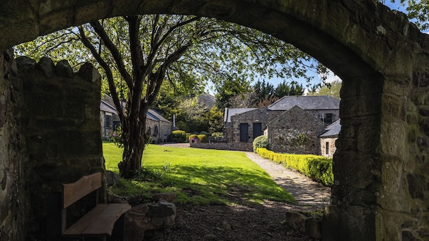 A stone archway leading to a stone building with a bench and a grassy area. - Timberwick Green (Wooler)