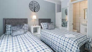 A bedroom with two beds, a nightstand with a lamp, a wall art piece, and a bathroom visible through an open door. - Flat 1A, Big Tree Holiday Flats (Paignton)