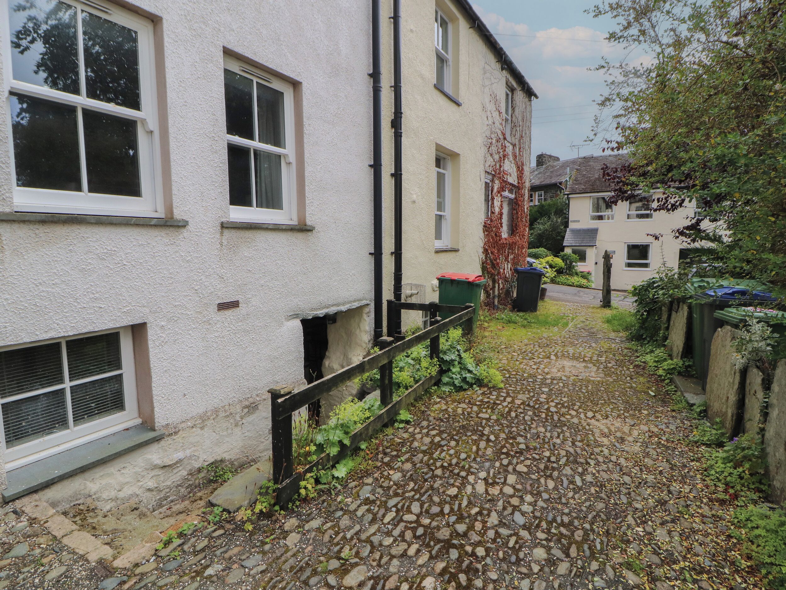 A narrow cobblestone pathway leading to a white building with multiple windows and a small garden area with a wooden fence.