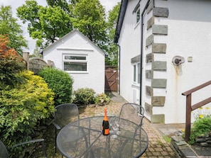 A white house with a stone exterior, a small garden with bushes and a tree, and a metal table with a bottle and glasses. - Hollens Cottage (Ambleside)