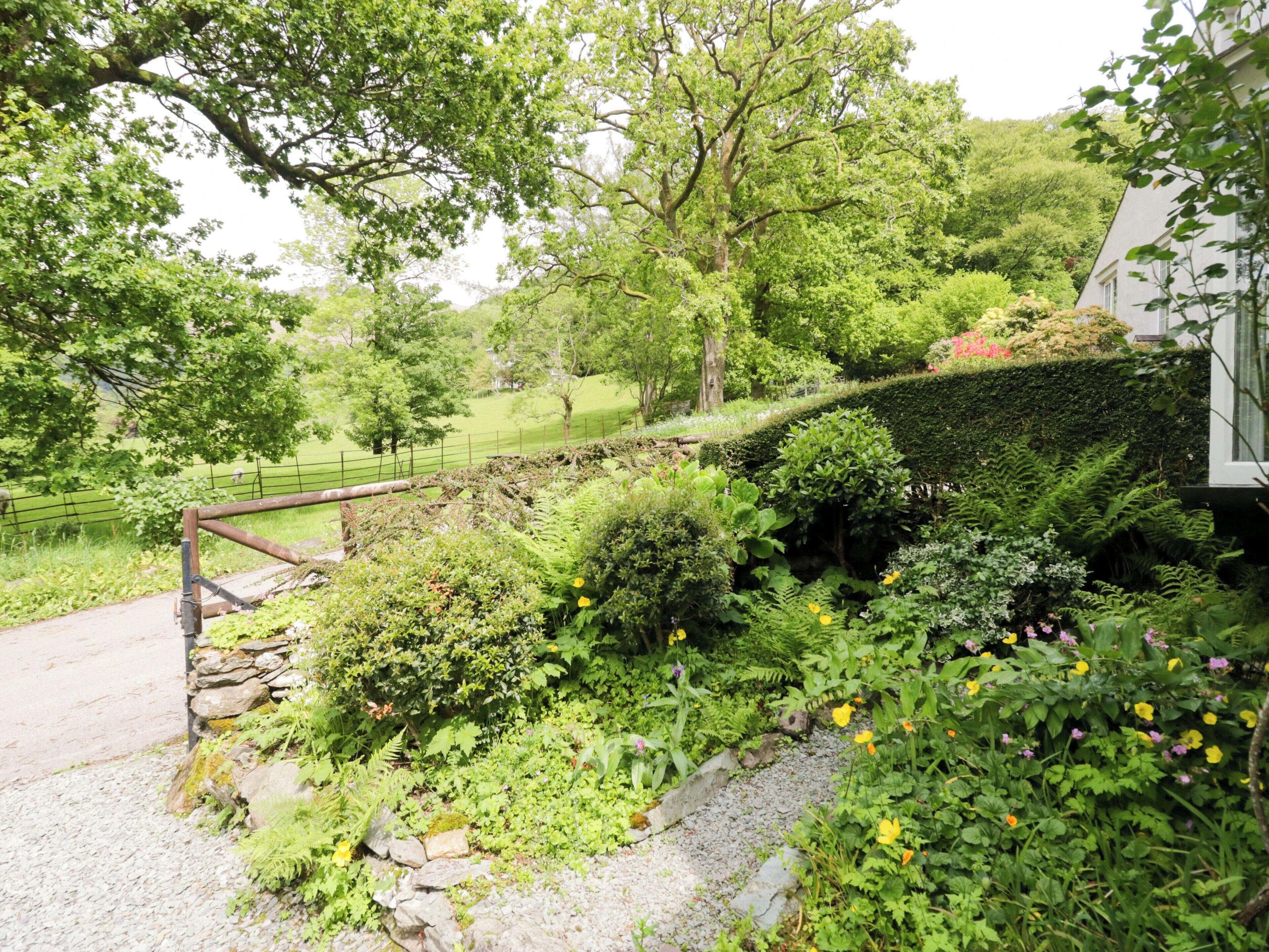 Um jardim com parede de pedra, caminho de cascalho e diversas plantas e flores.