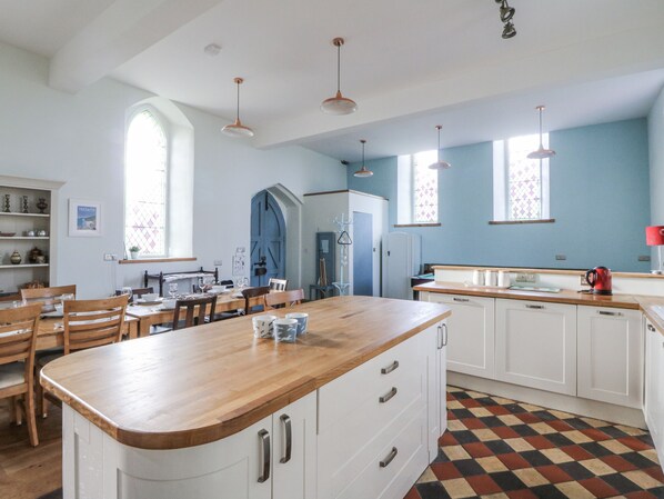 A spacious kitchen with a large island, white cabinets, and a checkered floor. There are pendant lights, a window with a view, and a dining area with wooden chairs and a table. - Church Llygad Y Dyffryn (Llandysul)