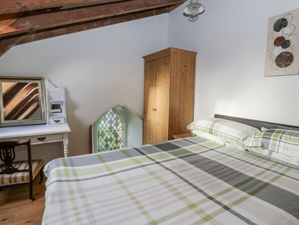 A bedroom with a bed, a wooden wardrobe, a desk with a mirror, and a window with a stained glass design.
