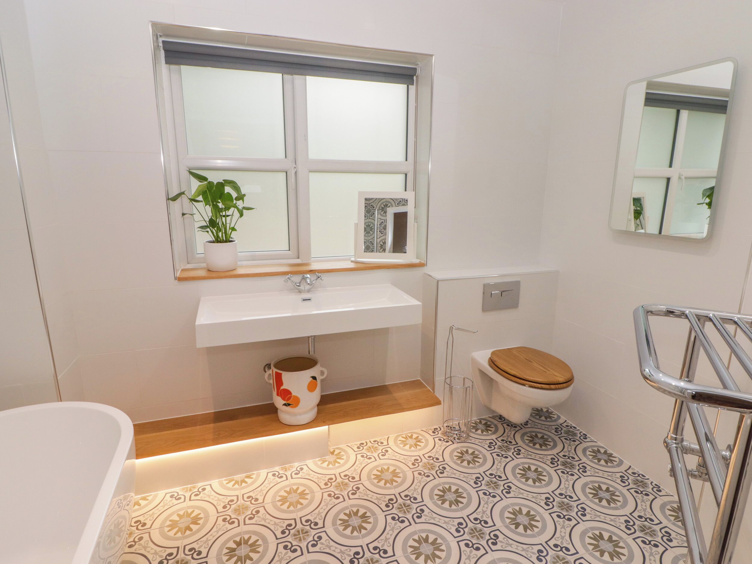 Un baño con lavamanos blanco, una ventana encima, inodoro con asiento de madera, bañera y baldosas de suelo estampadas.