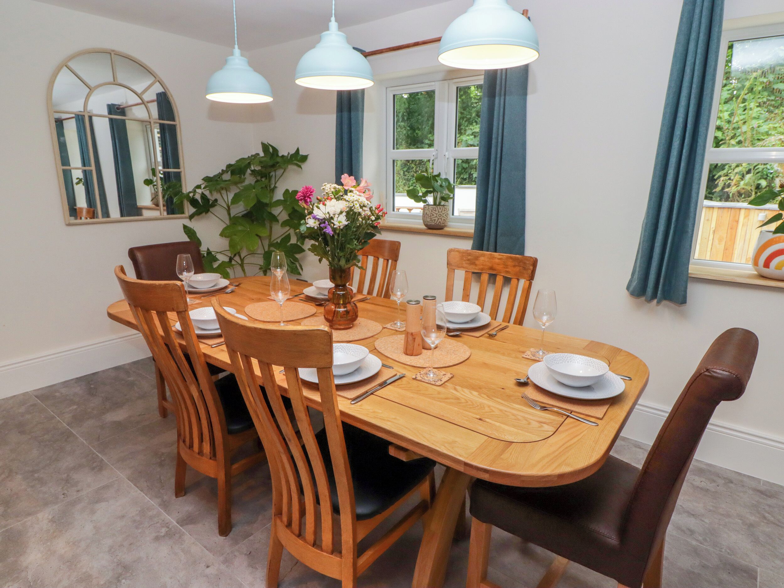 A dining room with a wooden table set for six, surrounded by wooden chairs. There are three hanging pendant lights above the table.