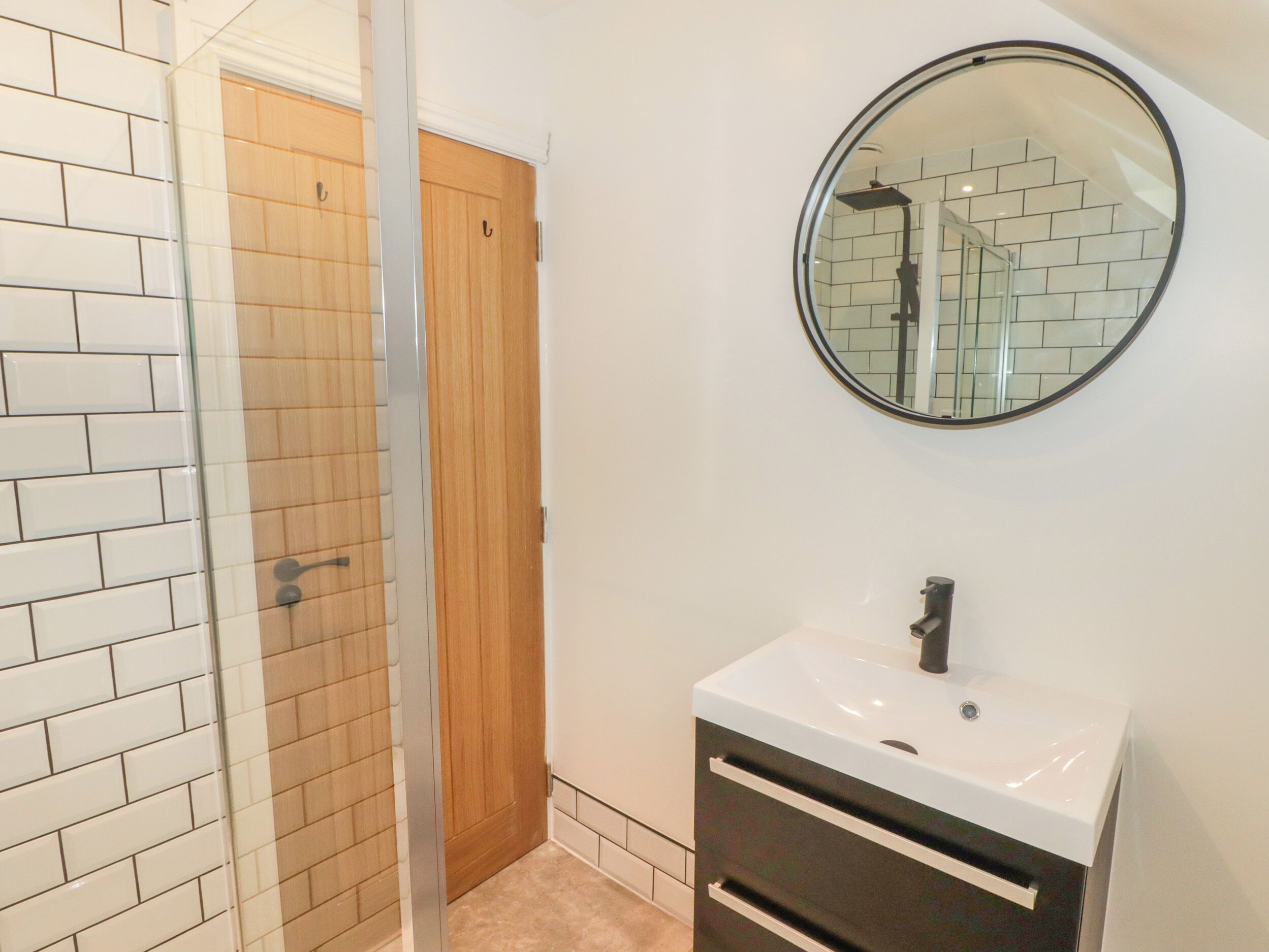A bathroom with a glass shower enclosure, a wooden door, a white sink with a mirror above it, and a tiled floor.
