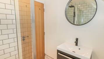 A bathroom with a glass shower enclosure, a wooden door, a white sink with a mirror above it, and a tiled floor.