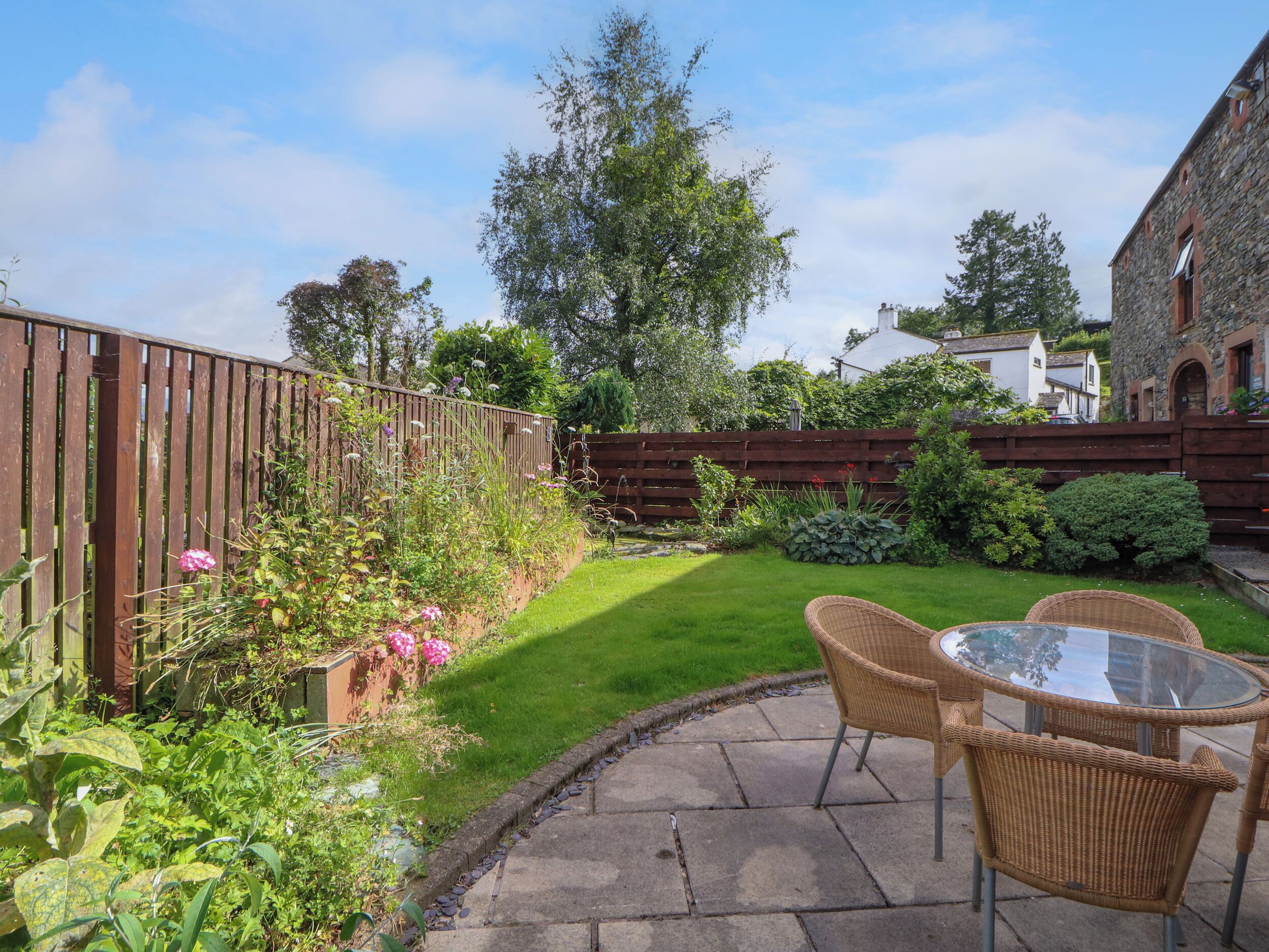 A patio with wicker chairs and a glass table, surrounded by a well-maintained garden with various plants and a wooden fence.