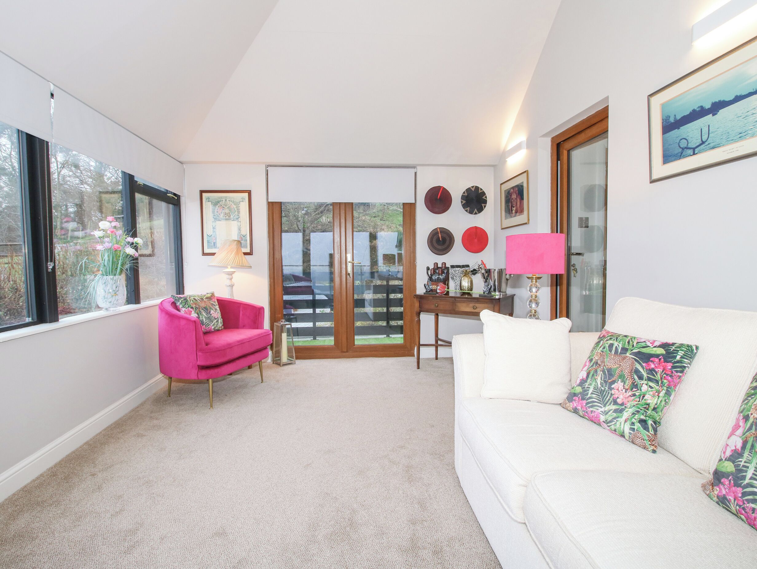 Una sala de estar con un sofá blanco, un sillón rosa y una mesa de centro. Hay una puerta que da a un balcón con vistas a árboles y una valla.