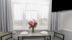 A dining area with a table set for two, a vase with flowers, and a window with curtains. - Flat 4, Big Tree Holiday Flats (Paignton)