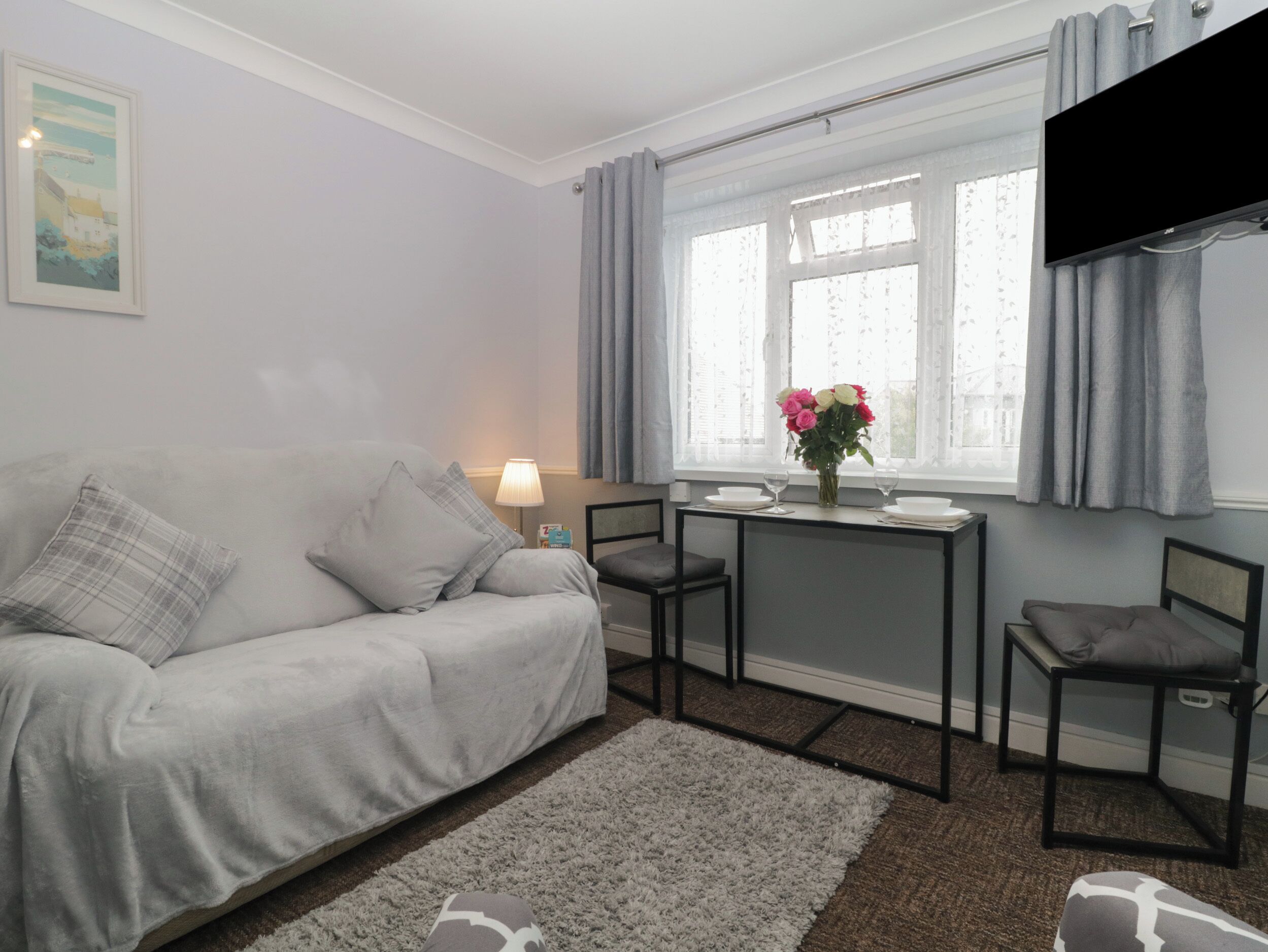 A modern living room with a grey sofa, a small table with a vase of flowers, and a TV mounted on the wall.