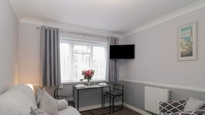 A compact hotel room with a bed, a small table with a vase of flowers, a TV mounted on the wall, and a patterned armchair. - Flat 4, Big Tree Holiday Flats (Paignton)