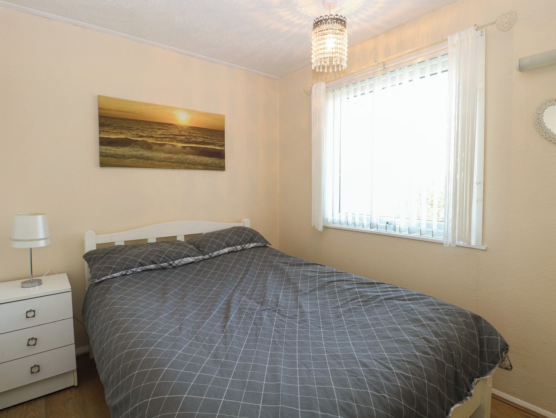 A bedroom with a bed, a nightstand, a lamp, a window with curtains, and a painting on the wall.