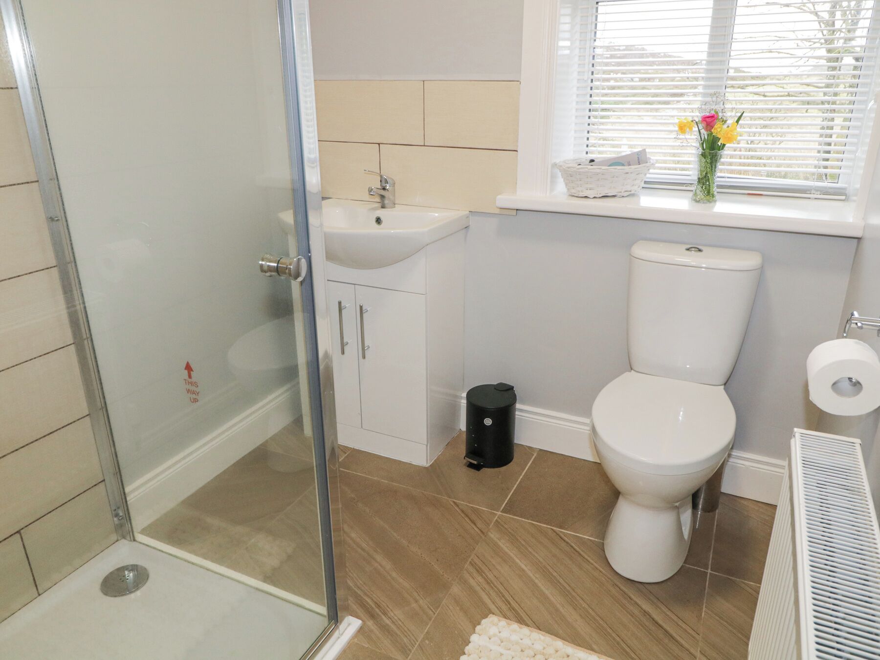 A bathroom with a glass shower enclosure, a sink, a toilet, and a window with a basket and flowers on the windowsill.