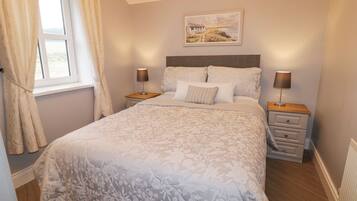 A neatly made bed with floral bedding, two bedside tables with lamps, a window with curtains, and a painting on the wall.