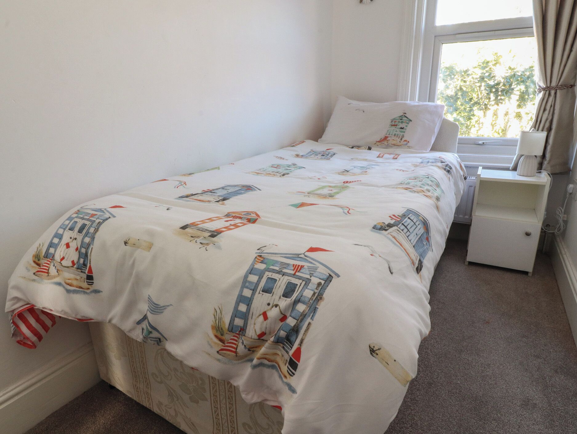 A single bed with a nautical-themed duvet cover, a bedside table with a lamp, and a window with curtains.
