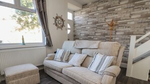 A living room with a beige sofa, matching ottoman, a geometric rug, a wooden wall, and a coat rack. - Harbour Mews (Lytham St. Anne's)