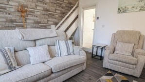 A living room with a grey sofa, striped pillows, a beige armchair, a geometric rug, and a wooden wall with a coat rack. - Harbour Mews (Lytham St. Anne's)