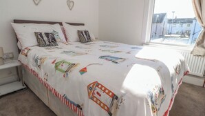 A bedroom with a bed, pillows, a bedside table, and a window with a view of buildings outside. - Harbour Mews (Lytham St. Anne's)