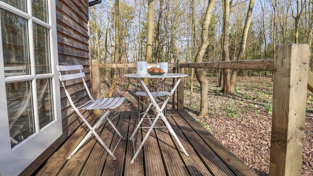 A wooden deck with a table and chairs, a window, and a view of a forest. - Woodlands Retreat Shepherd's Hut (Woodbridge)