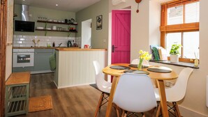 A compact kitchen with a dining area, a pink door, and a window with blinds. - Claire's Cottage (Bangor)