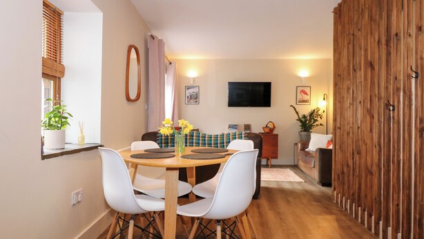 A dining area with a round wooden table, white chairs, a television on the wall, and a wooden partition. - Claire's Cottage (Bangor)