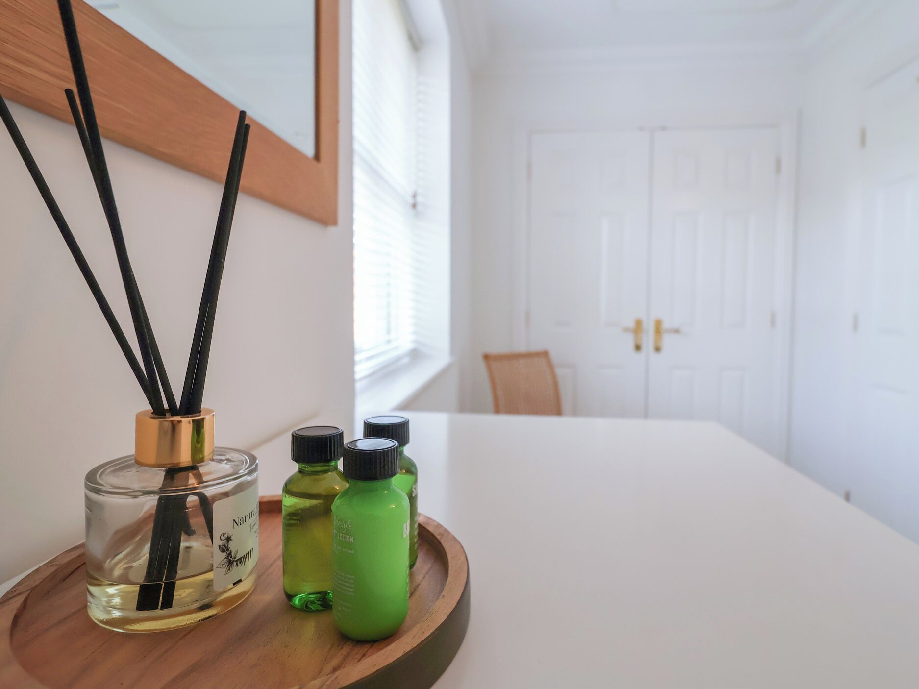 Une pièce avec une porte blanche, une chaise en bois et une table sur laquelle se trouve un plateau contenant un flacon de parfum, un flacon de lotion et un flacon d’huile essentielle.