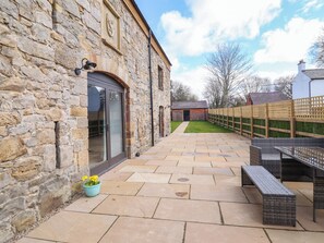 A stone building with a glass door, a paved patio with outdoor furniture, and a fenced garden area. - Threshers Barn (Oswestry)