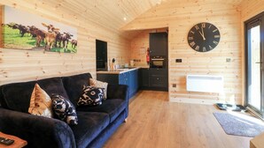 Ein gemütliches Blockhäuschen-Interieur mit Holzbalkendecke, einer schwarzen Couch mit gemusterten Kissen, einer großen Wanduhr und einer Küchenzeile.
