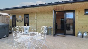 Ein Holzhaus mit überdachter Terrasse, inklusive Metalltisch und -stühlen, einem Whirlpool und einer Champagnerflasche.