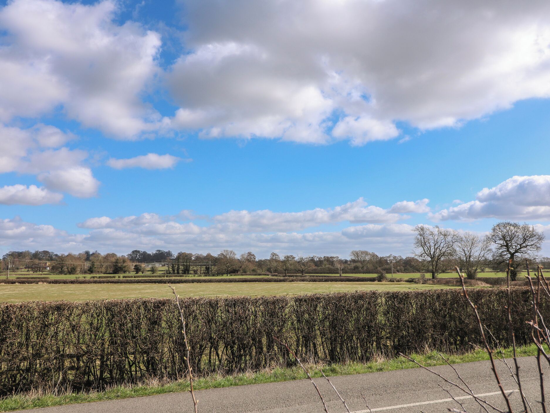 Un paysage rural avec une haie, une route et une ligne d’arbres à l’horizon, sous un ciel partiellement nuageux.