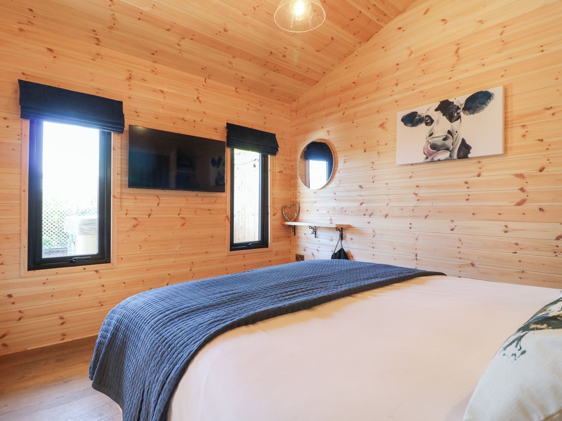 A wooden cabin bedroom with a bed, two windows, a mirror, and a painting on the wall.