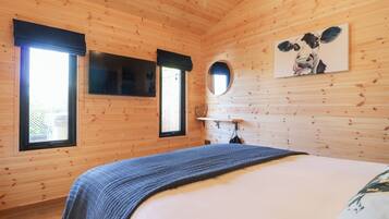A wooden cabin bedroom with a bed, two windows, a mirror, and a painting on the wall.