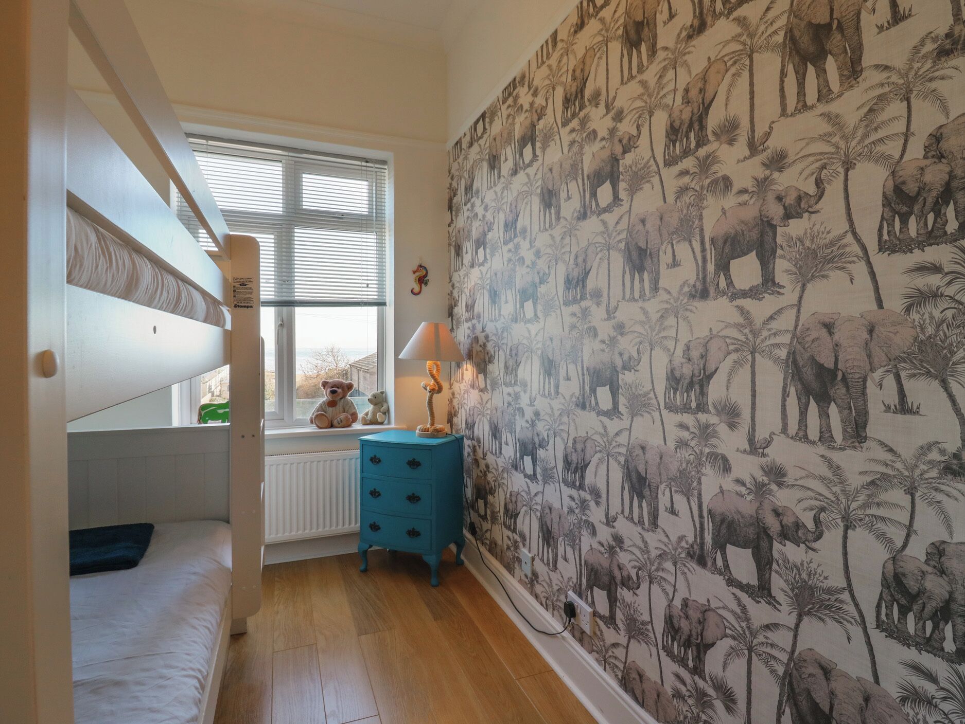 A child's bedroom with bunk beds, a window, a dresser, and a wall with elephant and palm tree wallpaper.
