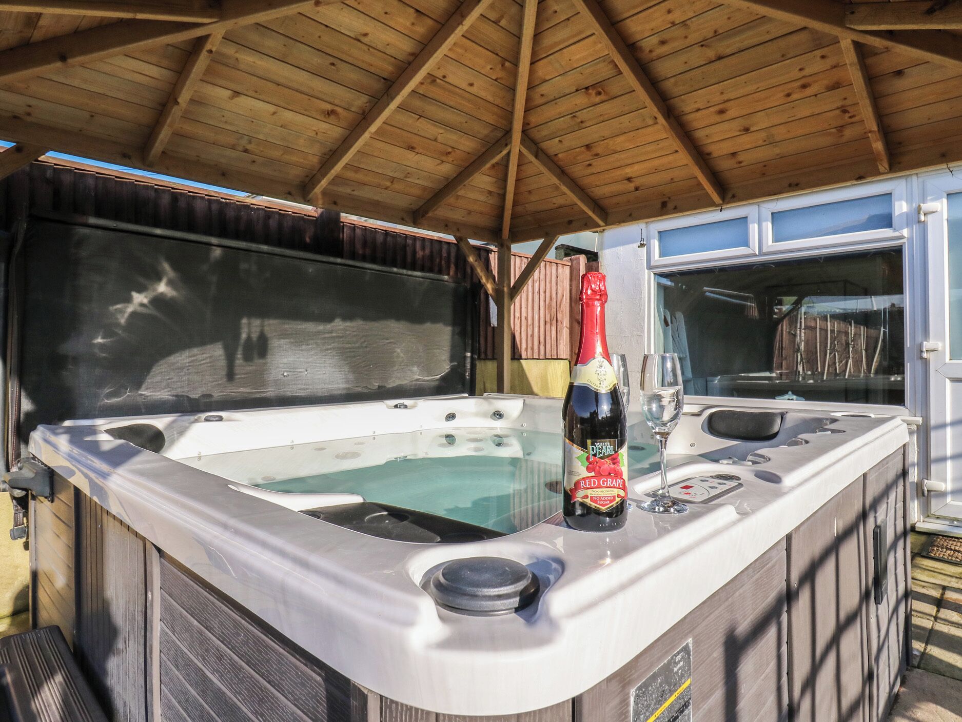 A hot tub with a bottle of wine and a glass, under a wooden roof structure.