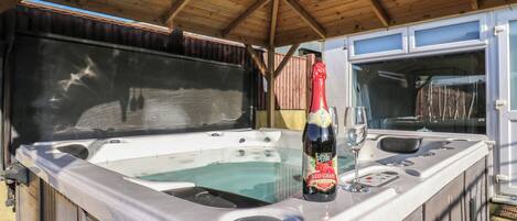 A hot tub with a bottle of wine and a glass, under a wooden roof structure.