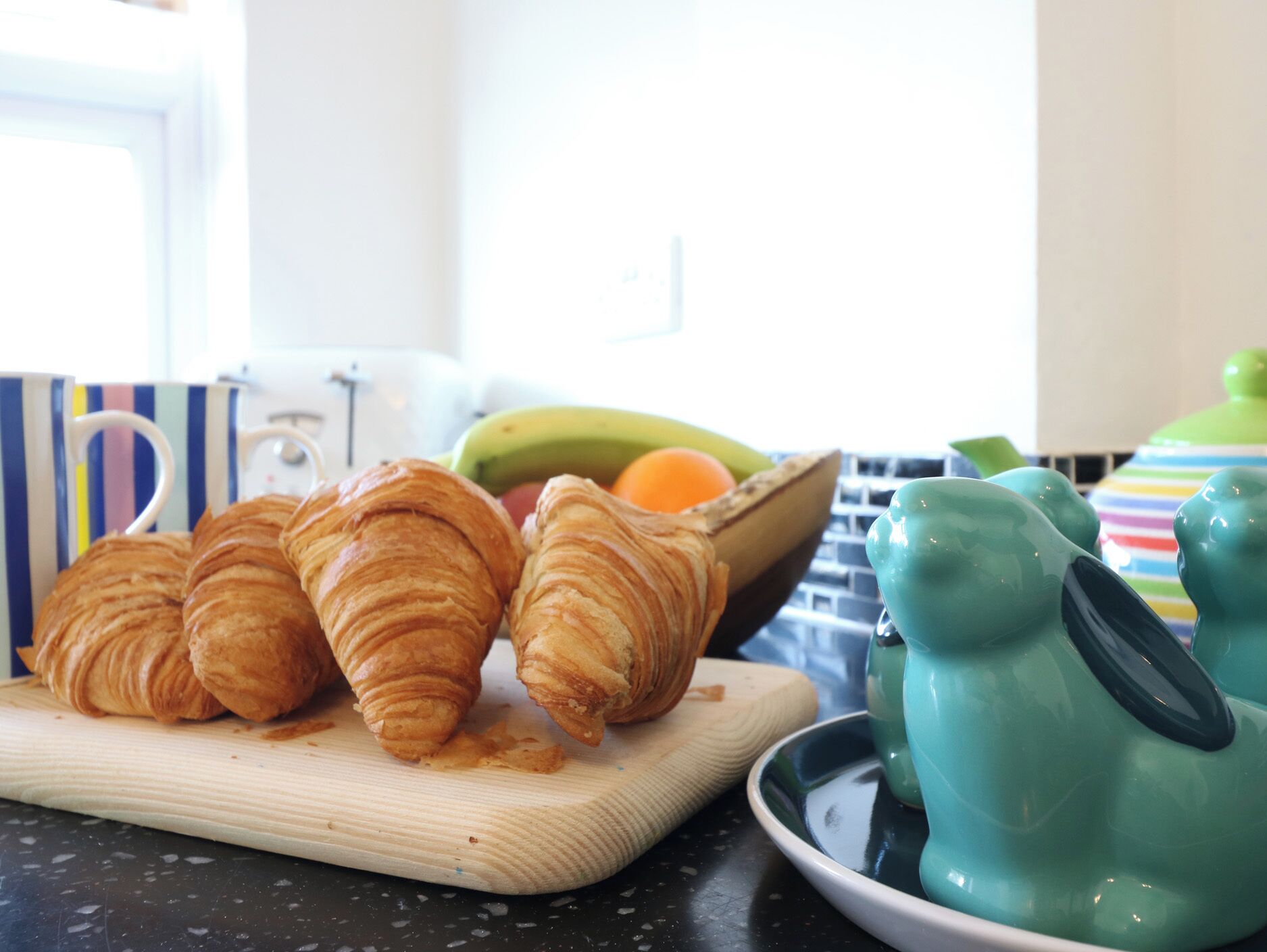 Croissants on a wooden cutting board, a bowl of fruit, and teal ceramic pitchers on a kitchen counter.